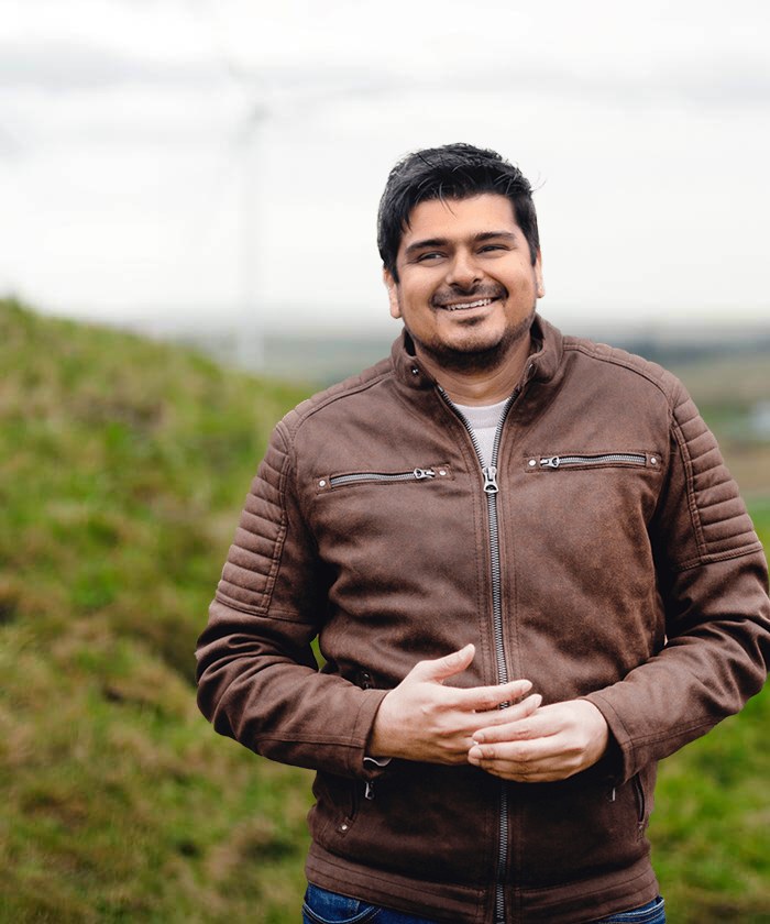 Ankit in een bruin leren jack op een dijk met windmolens