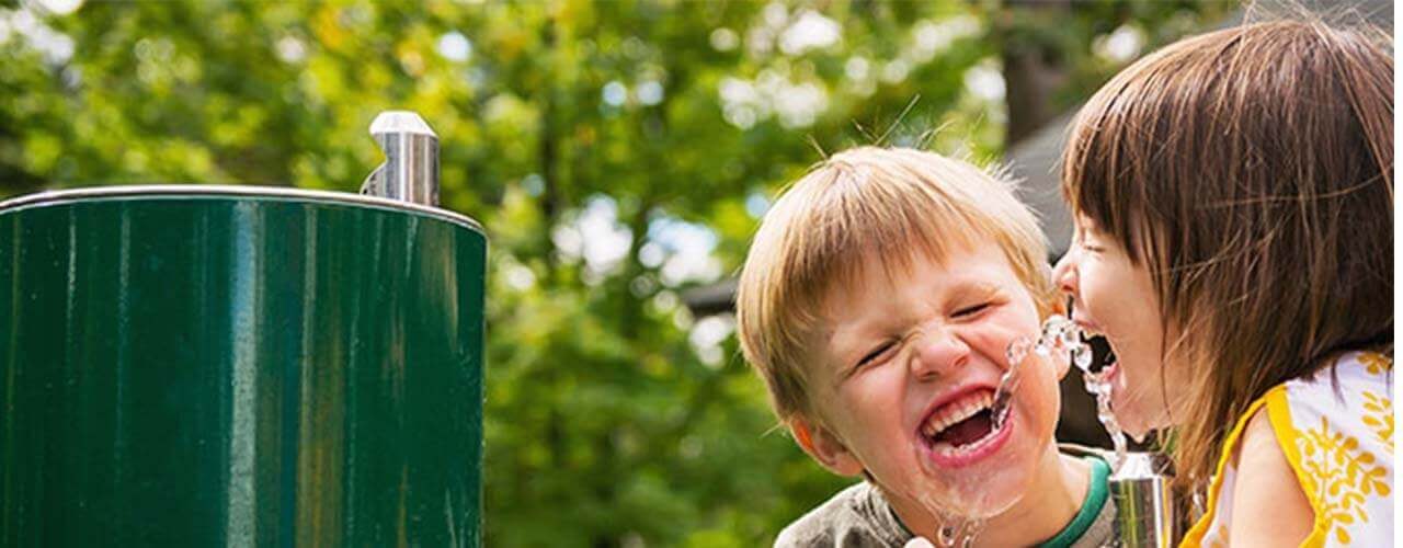 Twee kinderen drinken lachend uit een waterkraan in het park